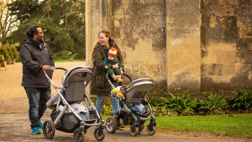 A family walking in the grounds at Christmas, Lacock Abbey, Wiltshire. They are pushing pushchairs and smiling. You can see glimpses of the abbey's front steps and Christmas trees in the background.